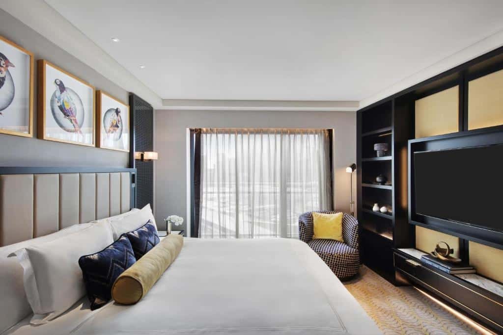 Quarto do The Star Grand Hotel and Residences. Do lado esquerdo uma mesa de casal, no fundo uma prateleira e um lustre. Do lado direito um painel com prateleiras, uma poltrona e um abajur. No fundo uma parede de vidro. 