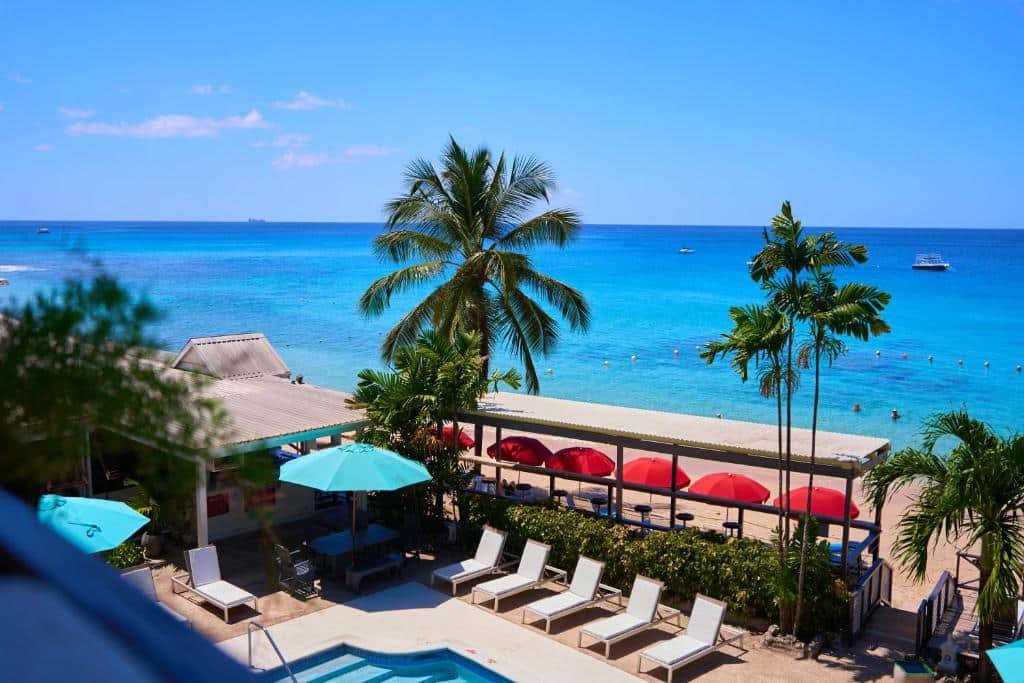 Parte do Tropical Sunset Beach que mostra parte da piscina aparecendo, em volta algumas espreguiçadeira, árvores verdes e guarda-sóis. Já ao fundo é possível ver o mar azul durante o dia. Imagem ilustrando post Hotéis em Barbados.
