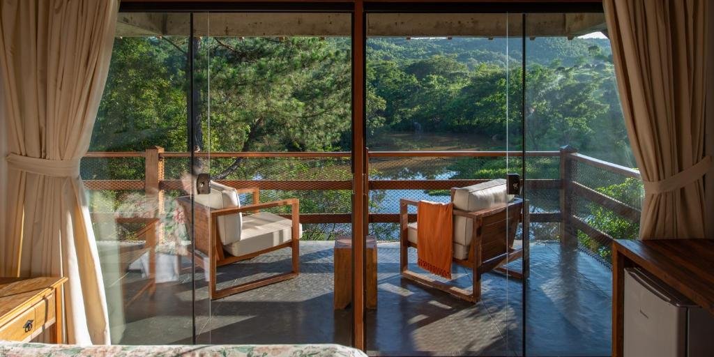 Varanda de chalé com cortinas bege, duas poltronas de madeira e estofado branco com parapeito de madeira e vista para um lago com árvores.