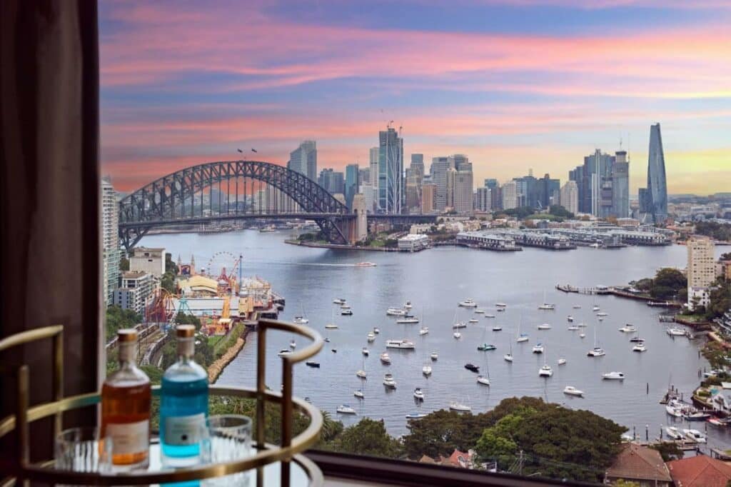 Vista da janela do View Sydney. Do lado esquerdo uma mesa com duas bebidas. A vista de um lago com barcos e uma ponte.