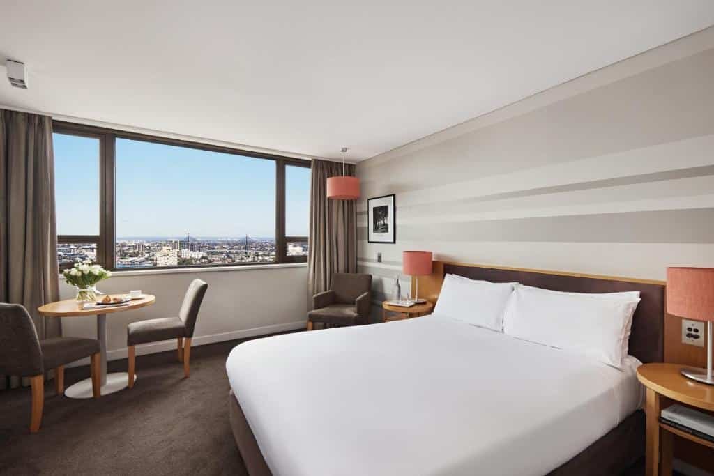 Quarto do View Sydney. Do lado direito uma cama de casal, de cada lado uma cômoda com um abajur, no fundo uma poltrona. Do lado esquerdo uma mesa com duas cadeiras, um vaso e atrás a janela.  Imagem para ilustrar post sobre hotéis em Sydney.