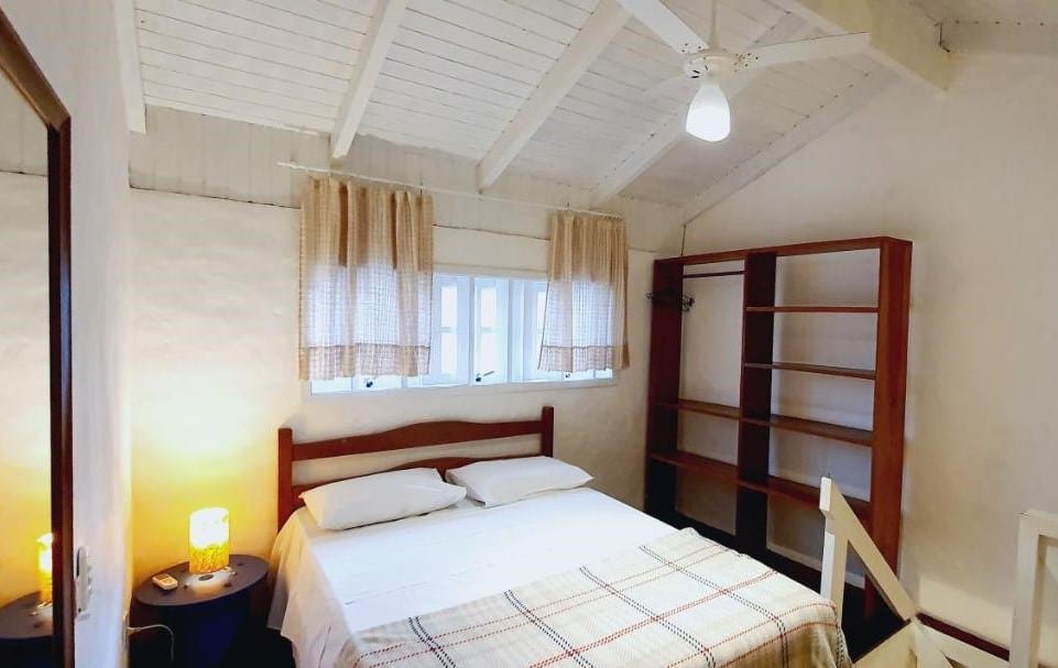 Quarto do Vilarejo Chalé. Uma cama de casal está no meio, abaixo de uma pequena janela. Ao seu lado esquerdo está uma pequena mesinha e um espelho, já do lado direito há um armário aberto.
