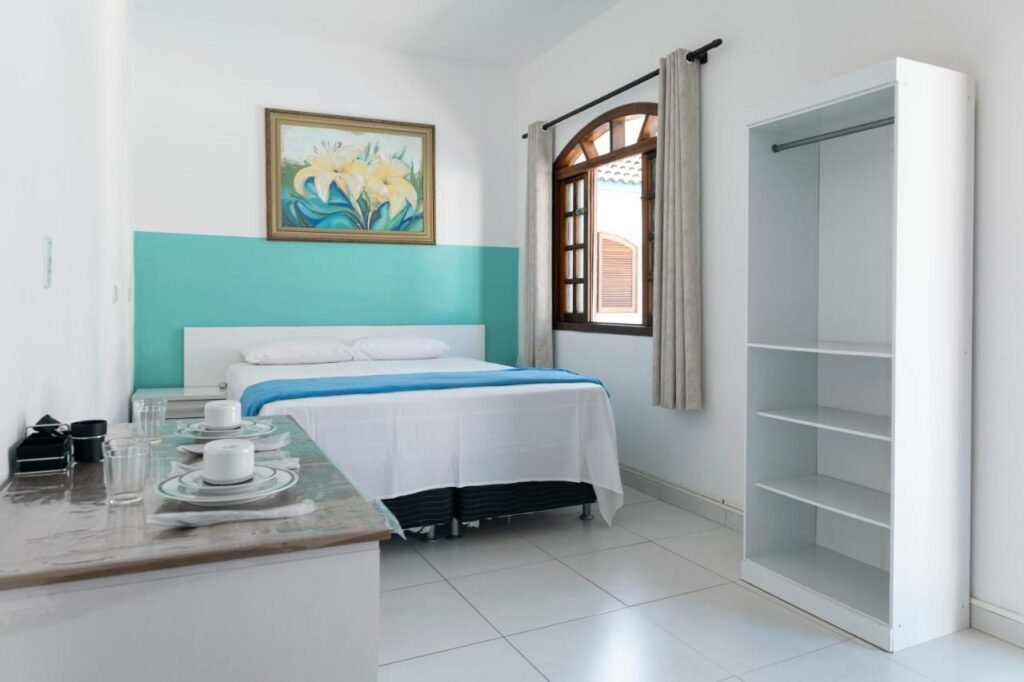 Quarto do Villa Salgada Maresias. Ao fundo há uma cama de casal cobrindo quase a parede toda e ao lado de uma janela. Mais para a frente, do lado direito, está um armário sem portas. Do lado esquerdo há um balcão com copos e xícaras.
.
