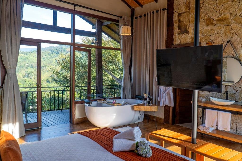 Imagem do quarto em Monte Mauá Chalés, que está ilustrando o post sobre airbnb em Visconde de Mauá. Há uma cama box de casal, e vemos o quarto como quem está deitado nela. Na frente há uma TV, fixa em uma coluna de ferro. Atrás há uma pia com espelho e uma banheira de hidromassagem do lado de uma janela de vidro com vista para as montanhas.