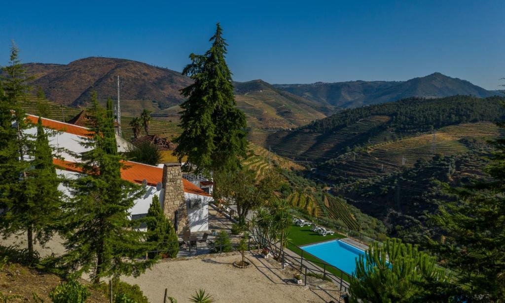 Vista da propriedade Quinta da Portela Douro com piscina do lado direito da imagem em meio as montanhas e do lado esquerdo a hospedagem em volta de árvores.