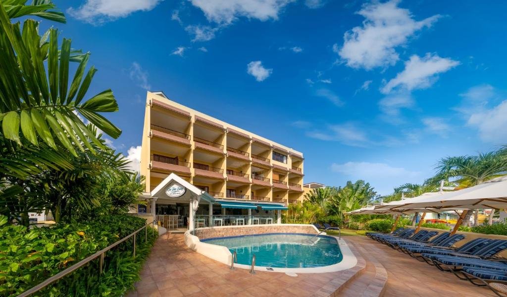 Parte externa do Yellow Bird Hotel que mostra uma piscina no centro e do lado direito algumas espreguiçadeiras e do lado direito algumas plantas verdes. Já ao fundo tem a construção do hotel com quatro andares. Imagem ilustrando post Hotéis em Barbados.